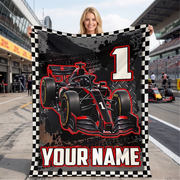 A woman stands on a racetrack holding a customizable black and red Formula 1 themed blanket with a checkered border. The blanket features a graphic of a black and red race car, a large number 1, and the text 'YOUR NAME'.