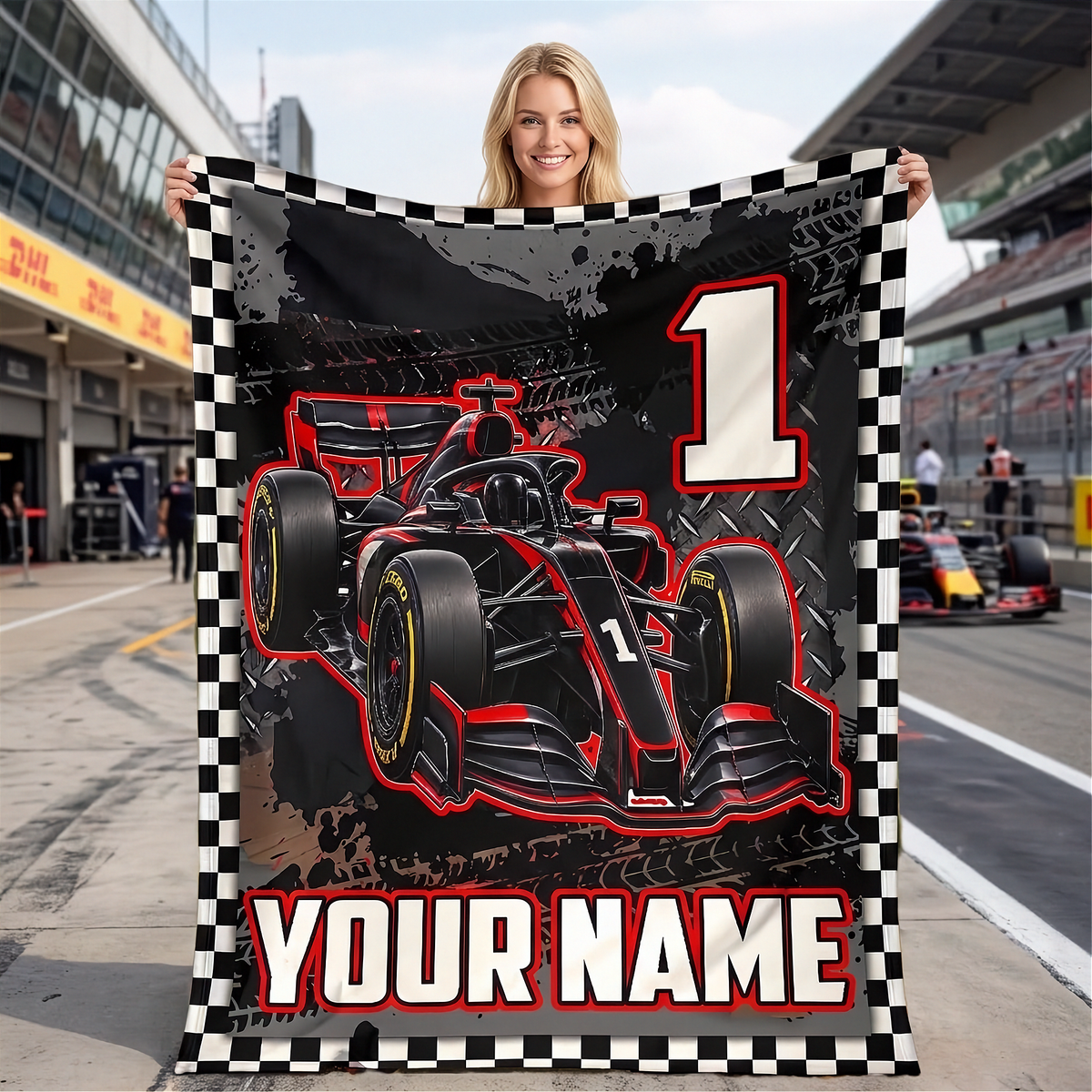 A woman stands on a racetrack holding a customizable black and red Formula 1 themed blanket with a checkered border. The blanket features a graphic of a black and red race car, a large number 1, and the text 'YOUR NAME'.