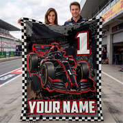 A man and a woman stand on a racetrack holding up a large customizable racing blanket. The blanket features a black and red Formula 1 race car, a large white number 1, a checkered border, and the text 'YOUR NAME' at the bottom.