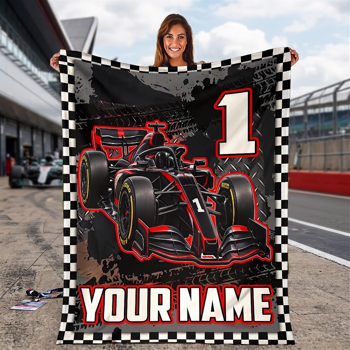 A woman stands on a racetrack holding a large customizable fleece blanket. The blanket features a black and red Formula 1 race car, a large white number 1, and a black-and-white checkered border, with the text 'YOUR NAME' at the bottom.