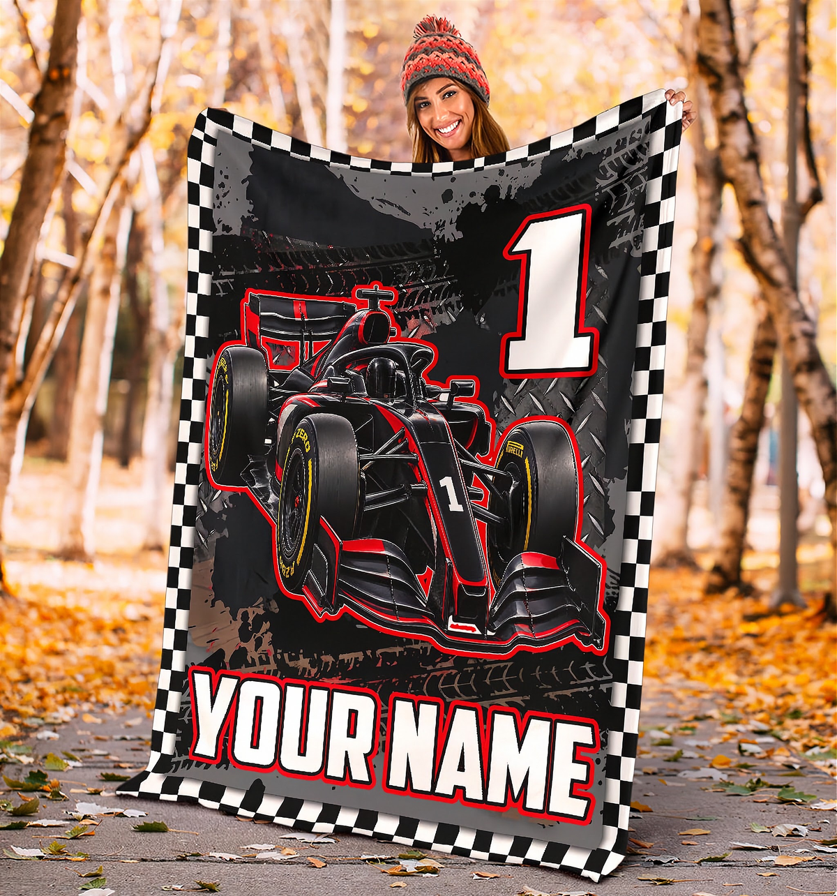 A fleece blanket featuring a red and black Formula 1 race car graphic, a checkered border, a large number 1, and the customizable text 'YOUR NAME', held up by a woman in a beanie in an autumn park setting.