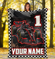 A woman holding up a custom printed fleece blanket featuring a black and red Formula 1 race car, a checkered border, a large number 1, and the text 'YOUR NAME'. The background is a blurred autumn forest with yellow leaves.