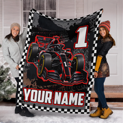 A custom printed fleece blanket featuring a black and red Formula 1 race car, a checkered border, a large number 1, and the text 'YOUR NAME'. Two young women are holding up the blanket in front of a white house door.