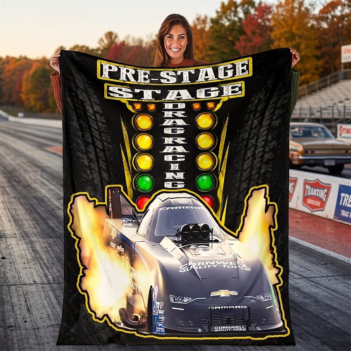 A woman holds up a large black fleece blanket featuring a drag racing graphic with the words 'PRE-STAGE DRAG RACING,' a black drag car with flames, and illuminated stoplights, set against a blurred drag strip background.