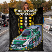 A woman stands in a racetrack holding up a large custom-printed blanket or poster featuring a green and white race car, a vertical row of colored lights, and the text 'PRE-STAGE STAGE YOUR NAME'.