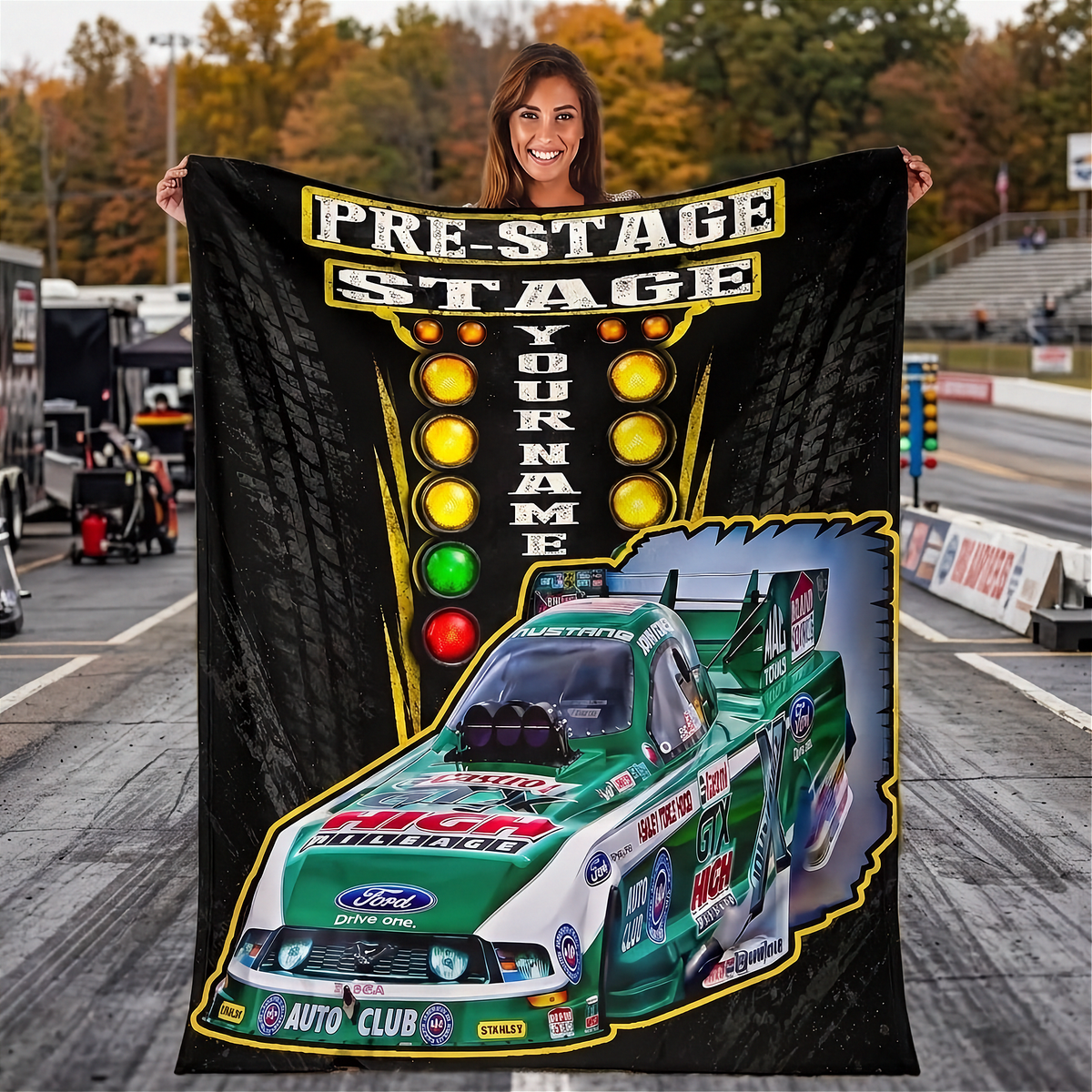 A woman stands in a racetrack holding up a large custom-printed blanket or poster featuring a green and white race car, a vertical row of colored lights, and the text 'PRE-STAGE STAGE YOUR NAME'.
