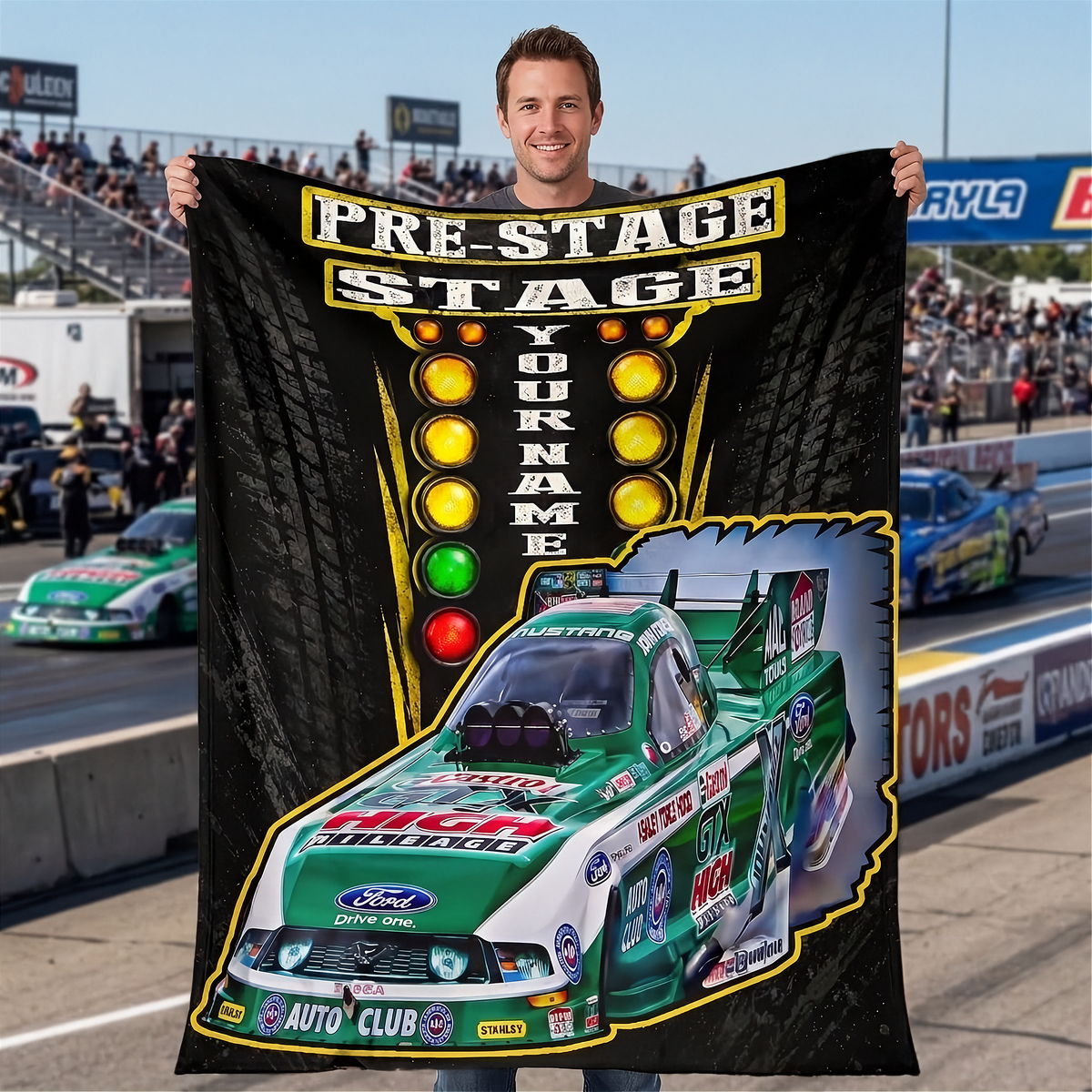 A man stands on a racetrack holding up a large custom-printed black blanket or towel featuring a green and white Ford race car, a light bar graphic, and the text 'PRE-STAGE STAGE YOUR NAME'.