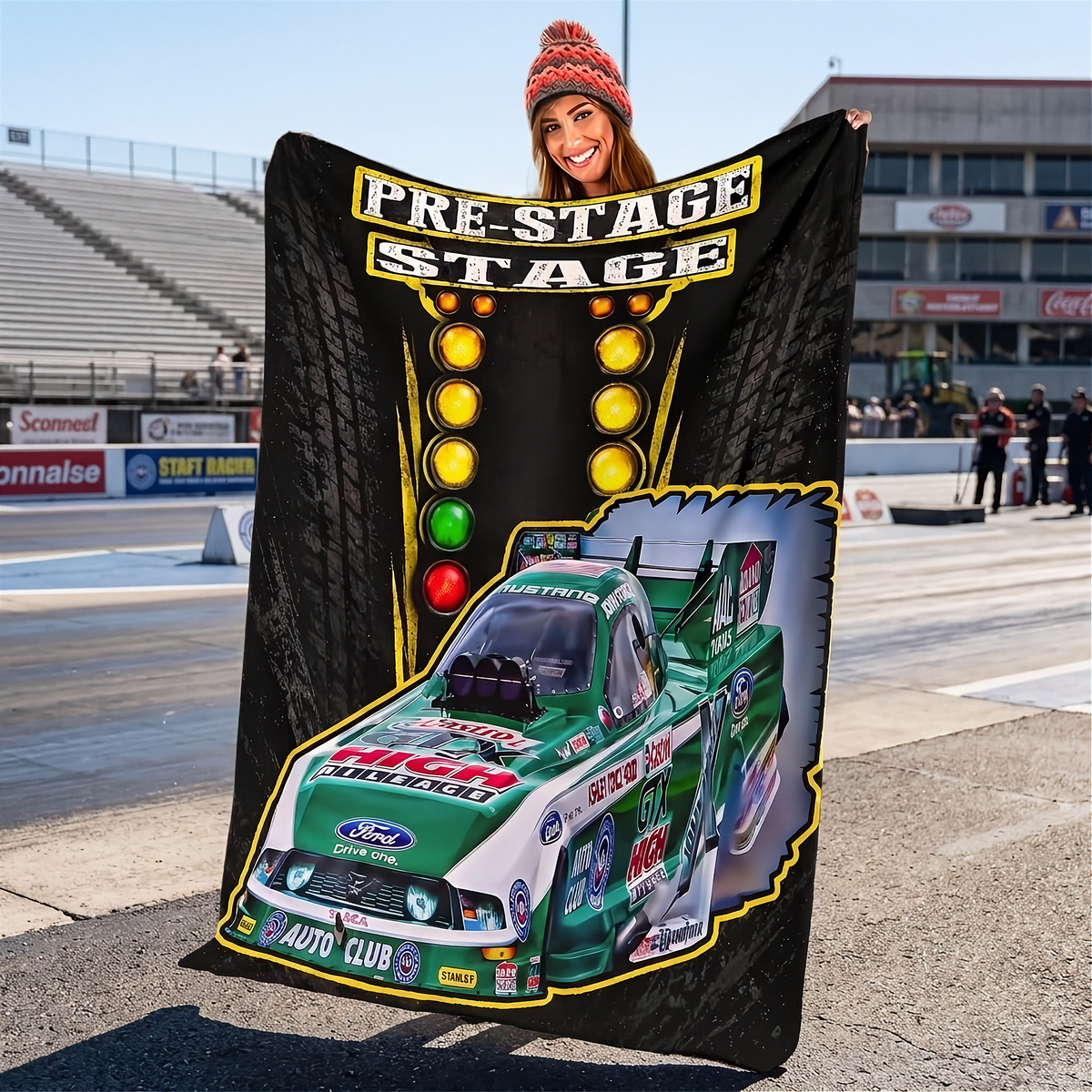 A woman wearing a red knit beanie holds up a large black fleece blanket or throw at an outdoor racetrack. The blanket features a racing design with the words 'PRE-STAGE' and 'STAGE' in yellow outlined letters, a vertical row of colored lights, and a green and white Ford race car graphic with sponsor logos.
