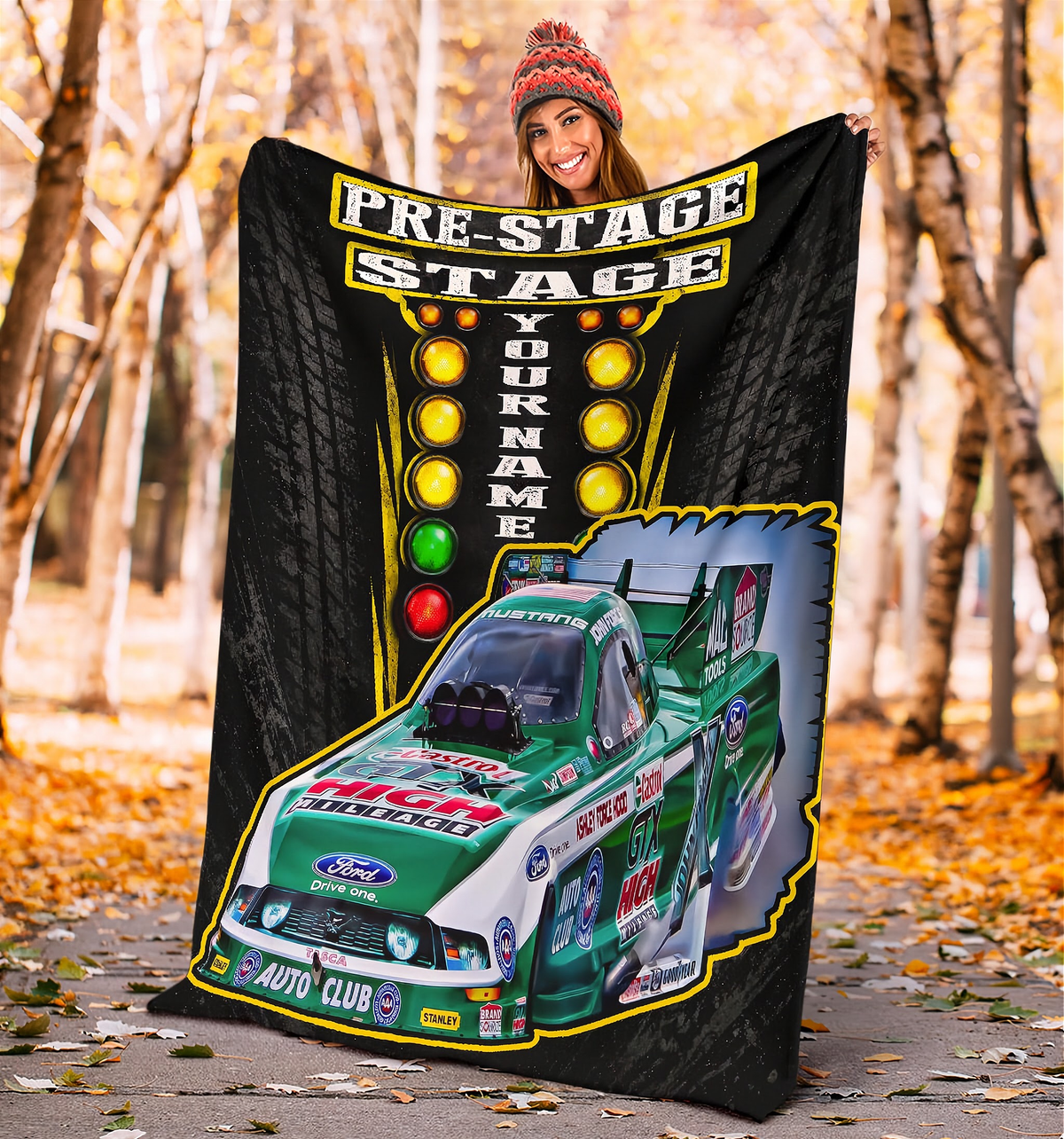 A woman stands outdoors in a wooded path holding up a large custom-printed blanket or poster. The product features a black background with tire tread graphics, a vertical marquee-style light bar, and a green and white racing truck graphic with sponsor logos.