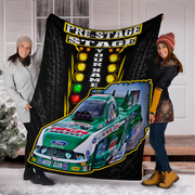 Two women holding up a large custom-printed black blanket or throw in front of a house door. The blanket features a green and white race car, the words 'PRE-STAGE STAGE' and 'YOUR NAME', and racing light graphics.