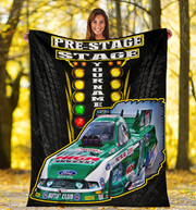 A woman stands outdoors in a wooded area holding up a large custom racing blanket. The blanket features a black background with tire tread graphics, a vertical light bar, the words 'PRE-STAGE STAGE' and 'YOUR NAME', and a green and white Ford race car graphic with sponsor logos.