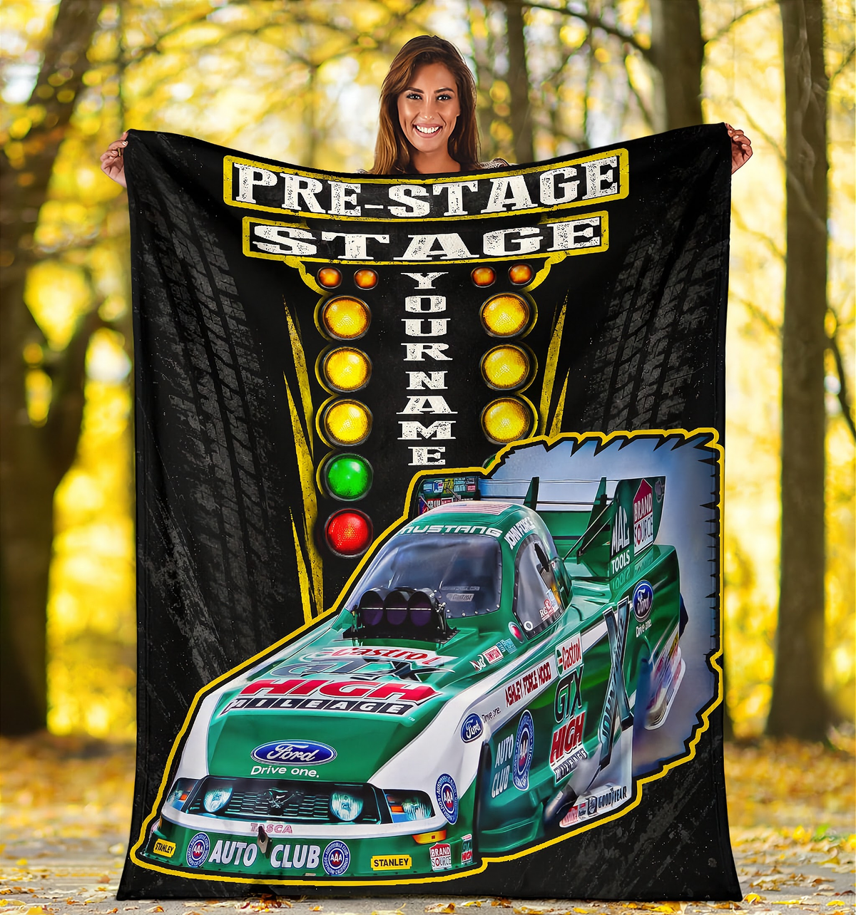 A woman stands outdoors in a wooded area holding up a large custom racing blanket. The blanket features a black background with tire tread graphics, a vertical light bar, the words 'PRE-STAGE STAGE' and 'YOUR NAME', and a green and white Ford race car graphic with sponsor logos.