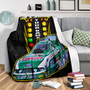 A custom printed fleece blanket draped over a gray sofa in a living room setting. The blanket features a green and white Ford race car with '2000 AUTO CLUB' and 'Ford' branding, alongside a black background with yellow, orange, and red traffic light graphics and the text 'YOUR NAME'.