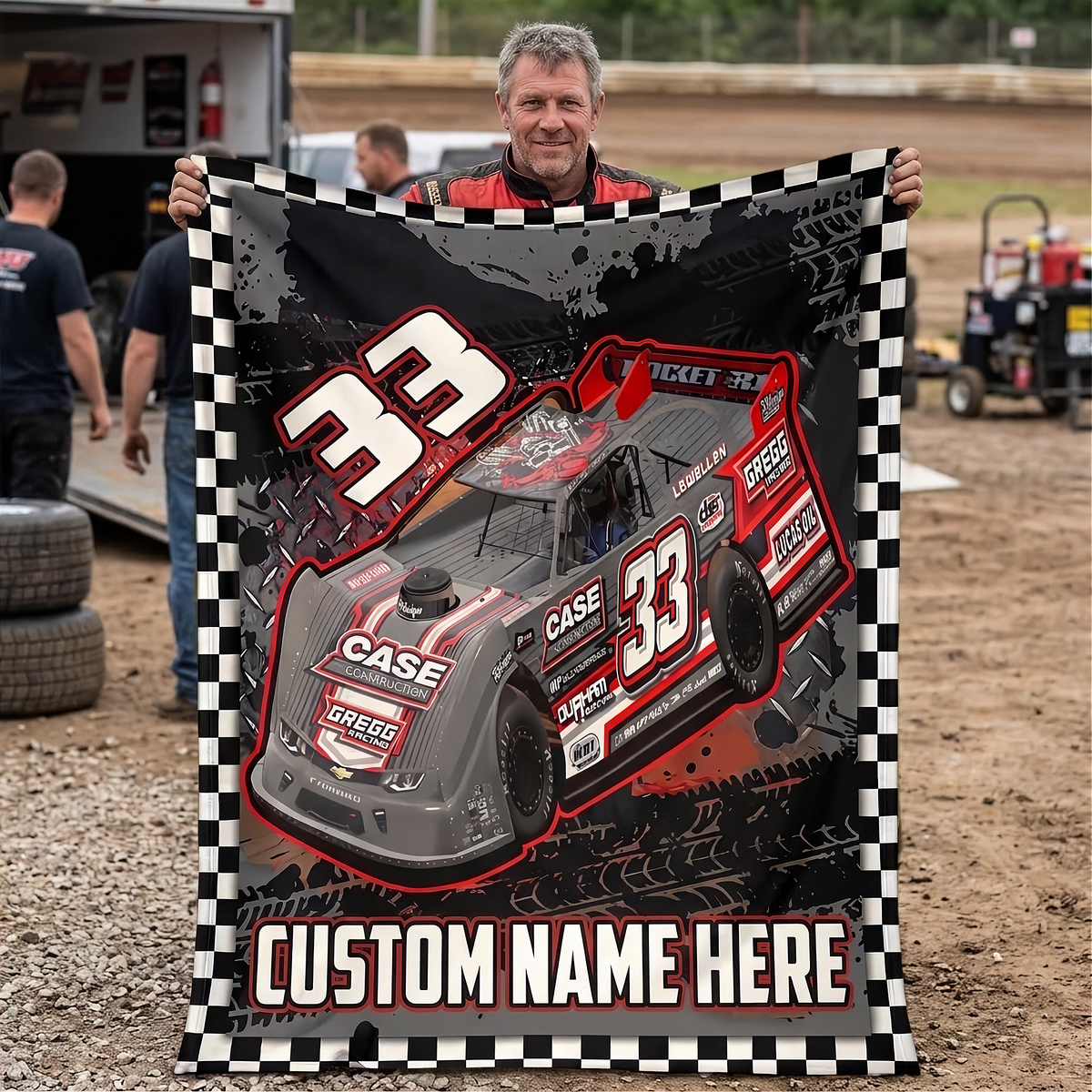 A man outdoors holds up a large custom race car blanket featuring a gray and red Late Model-style race car with the number 33, a black-and-white checkered border, and the text 'CUSTOM NAME HERE' at the bottom.