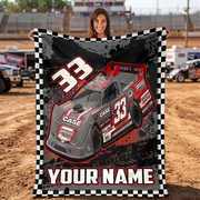 A woman stands in a dirt racing track holding up a large personalized blanket or poster featuring a red and gray race car with the number 33, a black-and-white checkered border, and the text 'YOUR NAME'.