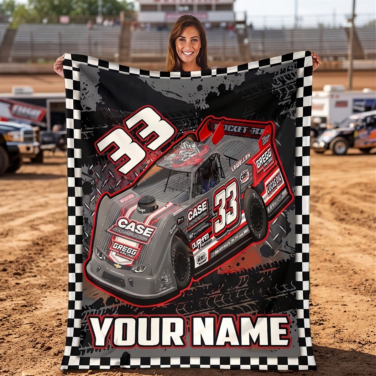 A woman stands in a dirt racing track holding up a large personalized blanket or poster featuring a red and gray race car with the number 33, a black-and-white checkered border, and the text 'YOUR NAME'.