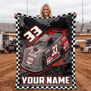 A woman stands in a dirt racing track holding up a large personalized blanket or poster featuring a gray and red race car with the number 33, a black-and-white checkered border, and the text 'YOUR NAME'.