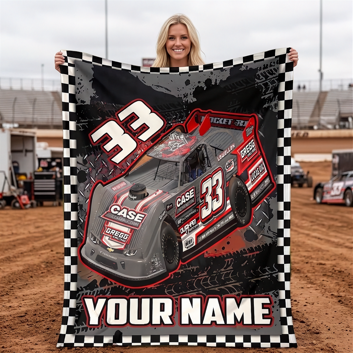 A woman stands in a dirt racing track holding up a large personalized blanket or poster featuring a gray and red race car with the number 33, a black-and-white checkered border, and the text 'YOUR NAME'.