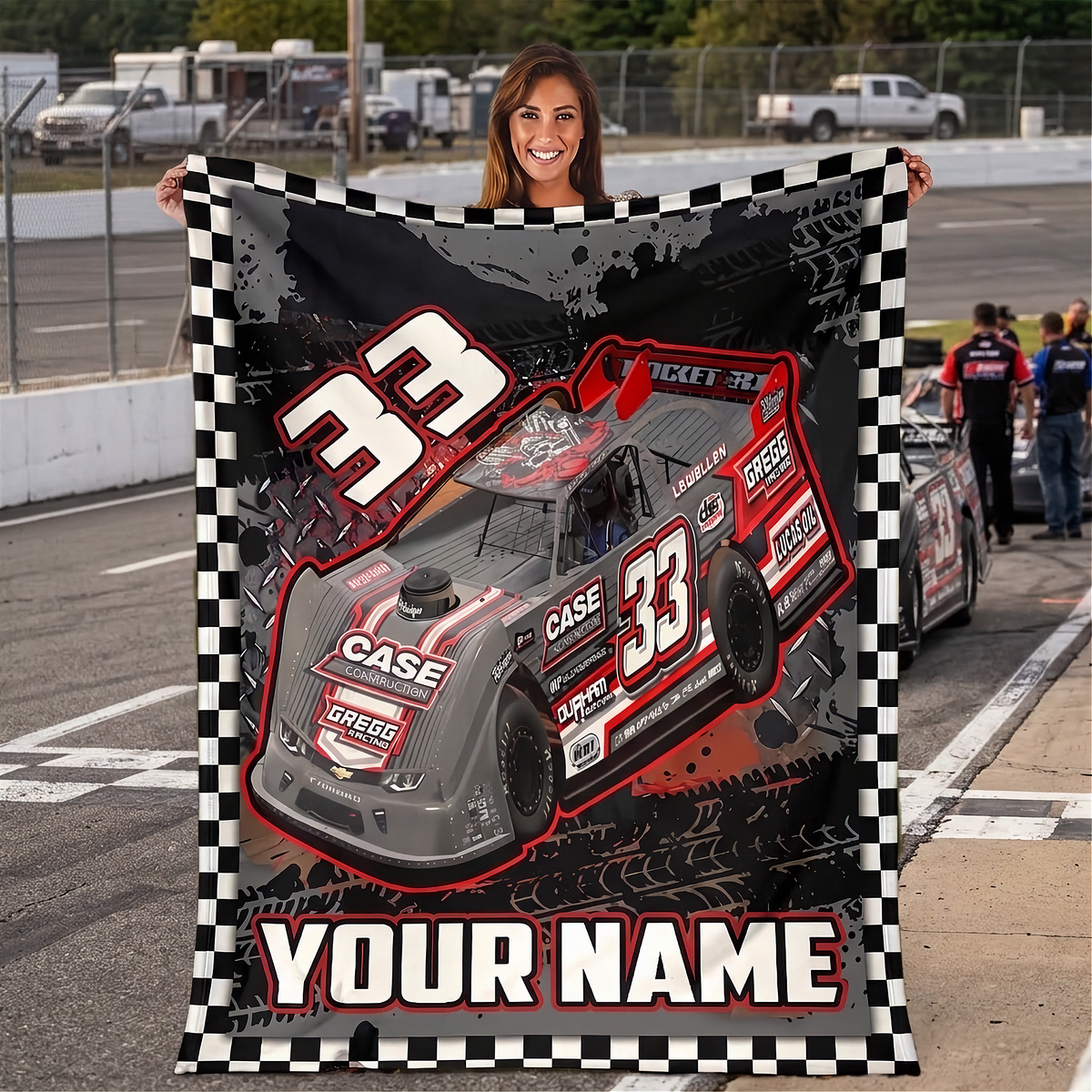 A woman stands on a racetrack holding up a large custom-printed blanket or poster featuring a gray and red race car with the number 33, a black-and-white checkered border, and the text 'YOUR NAME' at the bottom. A race car is visible in the background on the track.