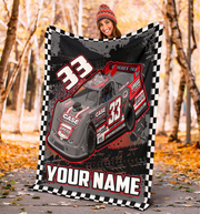 A person wearing a red knit beanie holds up a large custom race car blanket featuring a gray and red Late Model race car with the number 33, a black-and-white checkered border, and the text 'YOUR NAME' at the bottom. The background is an outdoor autumn forest path with fallen leaves and tree trunks.
