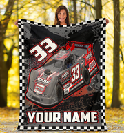 A woman outdoors in a wooded setting holds up a large custom race car blanket. The blanket features a black and white checkered border, a gray and red race car with the number 33, and the text 'YOUR NAME' at the bottom.
