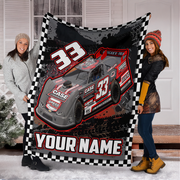 Two women standing outdoors in front of a house door holding up a large personalized racing blanket. The blanket features a black and white checkered border, a red and gray late model race car with the number 33, and the text 'YOUR NAME'.