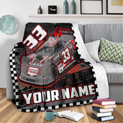 A custom racing-themed throw blanket draped over a gray sofa in a living room setting. The blanket features a graphic of a gray and red late model race car with the number 33, sponsor logos, a black-and-white checkered border, and the customizable text 'YOUR NAME'.