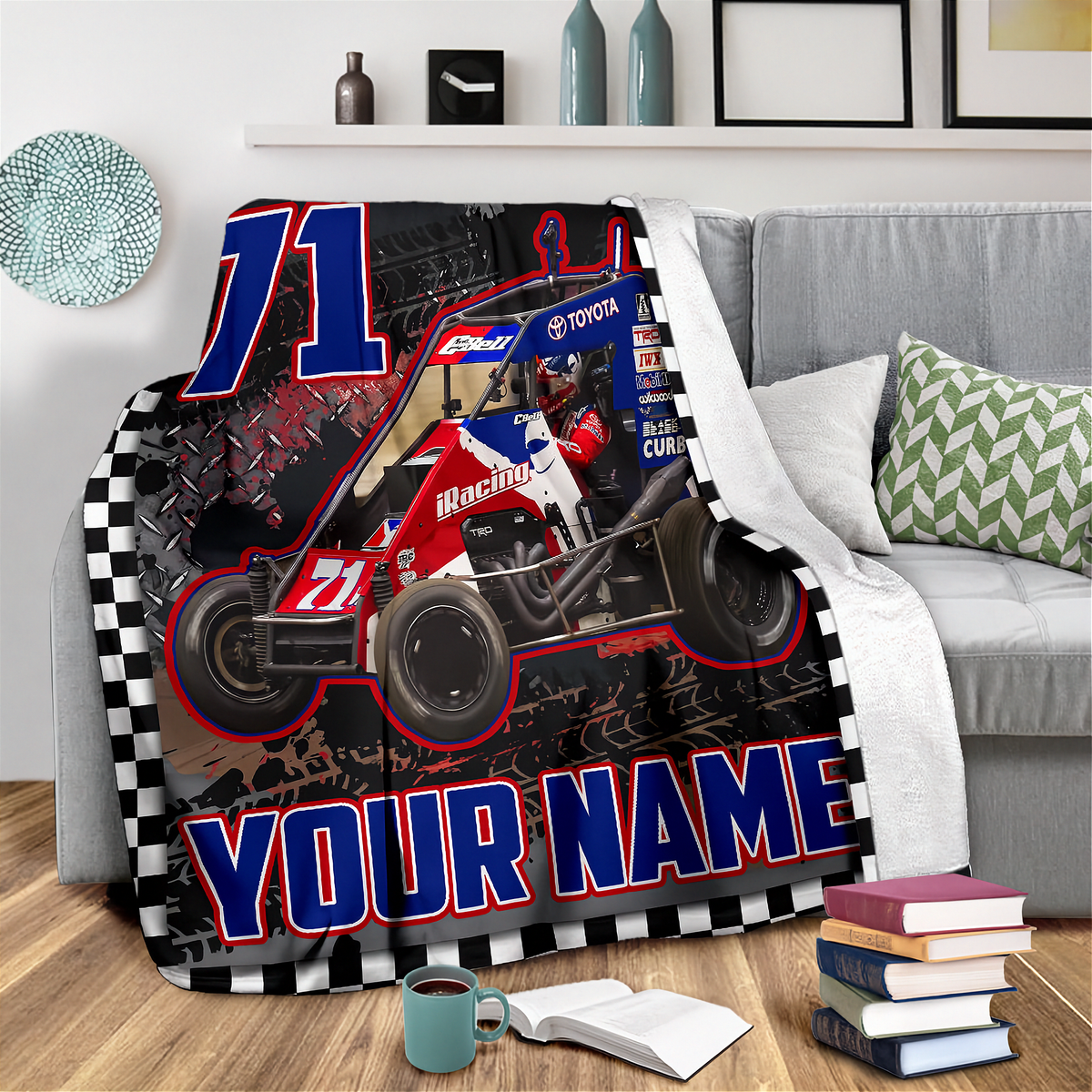 A custom printed racing blanket draped over a gray sofa in a living room setting. The blanket features a red and blue midget race car with the number 71, a checkered border, and 'YOUR NAME' printed in bold blue letters at the bottom.