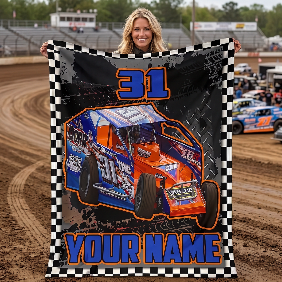 A woman stands on a dirt racetrack holding up a large custom-printed blanket or poster featuring a blue and orange race car with the number 31, a black-and-white checkered border, and the text 'YOUR NAME'.