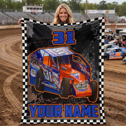 A woman stands on a dirt racetrack holding up a large custom-printed blanket or poster featuring a blue and orange race car with the number 31, a black-and-white checkered border, and the text 'YOUR NAME'.