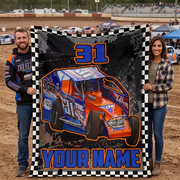 A man and a woman standing on a dirt racetrack holding a large custom-printed blanket. The blanket features a blue and orange race car, the number 31, tire tracks, and the text 'YOUR NAME'.
