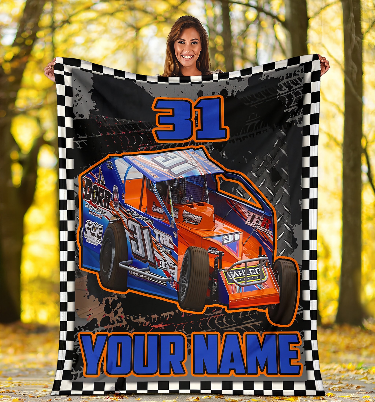 A woman holds up a large personalized blanket featuring a blue and orange race car with the number 31, a black-and-white checkered border, and the text 'YOUR NAME' at the bottom, set against a blurred autumn forest background.