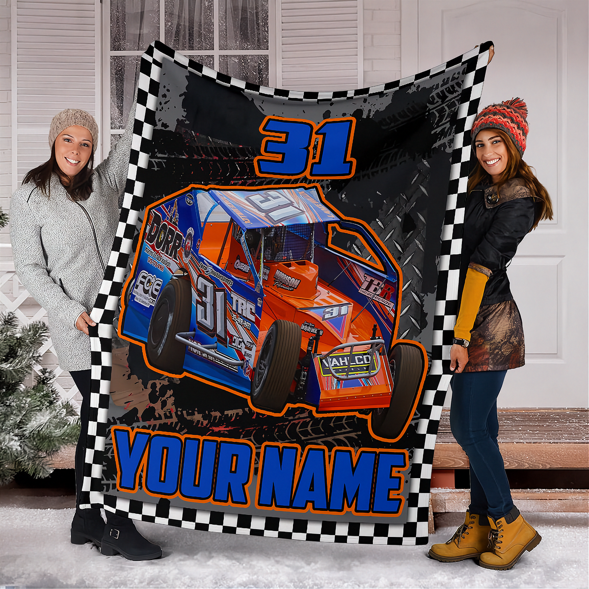 Two women holding up a large custom race car blanket in front of a snowy porch. The blanket features a black and white checkered border, a blue and orange race car graphic, the number '31', and the text 'YOUR NAME'.