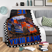 A custom printed racing blanket draped over a gray sofa in a living room setting. The blanket features a blue and orange race car with the number 31, a black and white checkered border, and the customizable text 'YOUR NAME'.