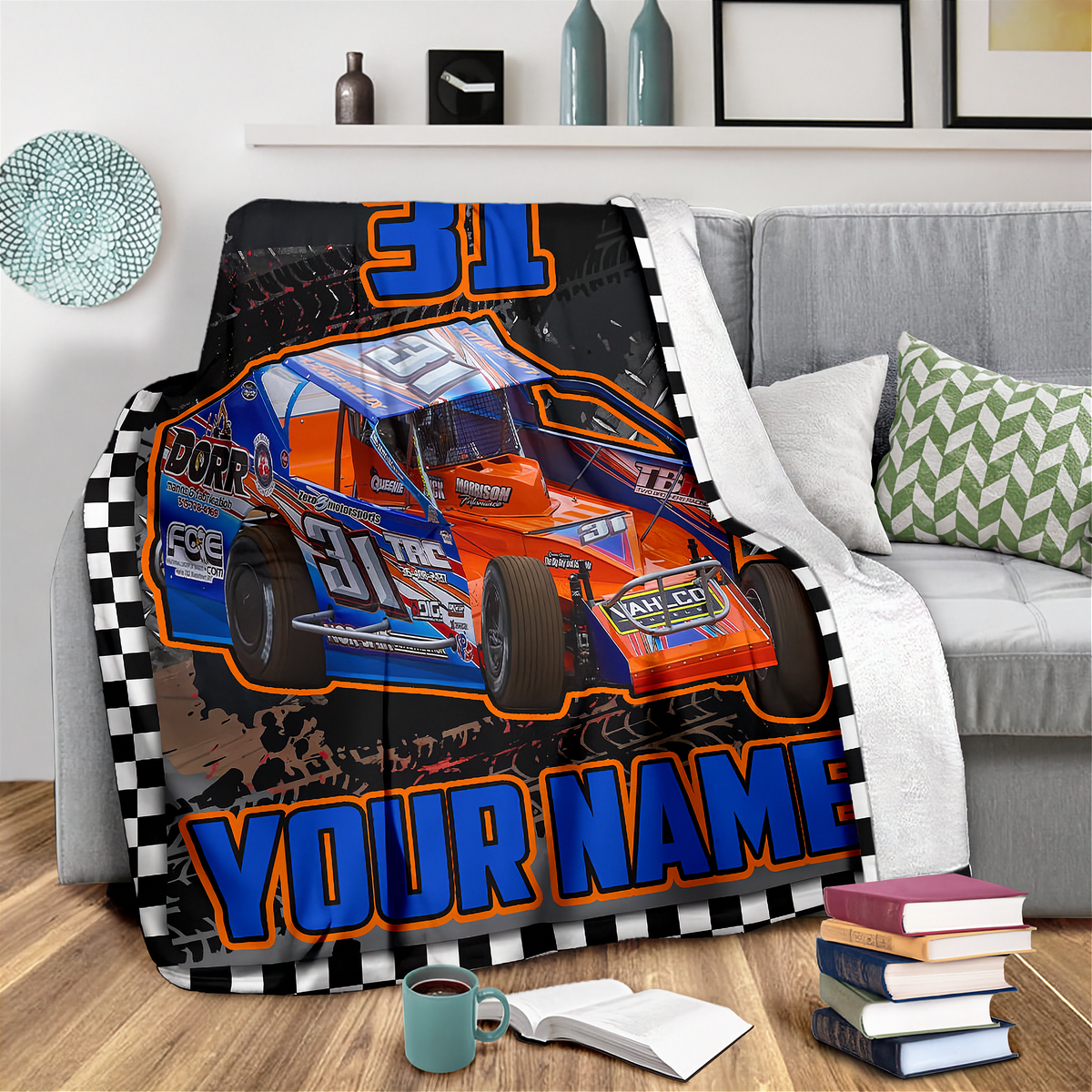 A custom printed racing blanket draped over a gray sofa in a living room setting. The blanket features a blue and orange race car with the number 31, a black and white checkered border, and the customizable text 'YOUR NAME'.