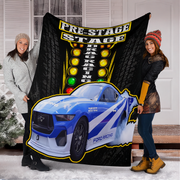 Two women holding up a large black fleece blanket featuring a blue and white Ford Mustang race car, yellow 'PRE-STAGE' and 'DRAG RACING' text, and a light-topped track backdrop, set in front of a white house door with snow on the ground.