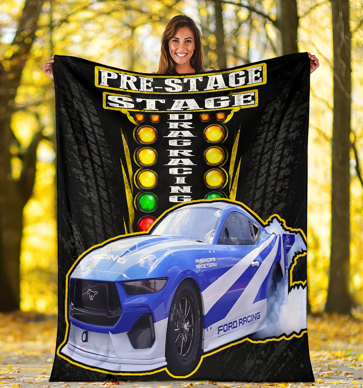 A woman stands outdoors in a wooded area holding up a large printed blanket or tapestry. The product features a blue and white Ford mustang racing car with yellow, white, and black graphics, plus a vertical race-light style design and the words 'PRE-STAGE STAGE' and 'DRAG RACING'.