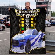 A man stands in a racetrack parking area holding up a large black printed blanket or poster featuring a blue Ford racing car and the words 'PRE-STAGE DRAG RACING' with illuminated race lights. The background shows race cars, track barriers, and grandstands.