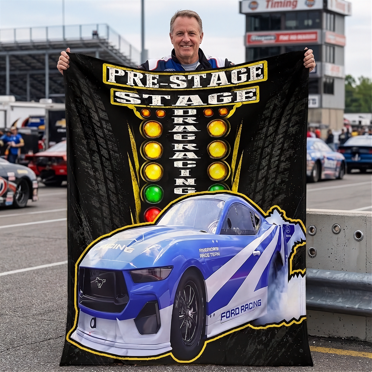 A man stands in a racetrack parking area holding up a large black printed blanket or poster featuring a blue Ford racing car and the words 'PRE-STAGE DRAG RACING' with illuminated race lights. The background shows race cars, track barriers, and grandstands.