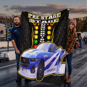A man and a woman stand on a drag strip holding up a large custom-printed blanket or tapestry. The blanket features a black background with a tire tread pattern, 'PRE-STAGE DRAG RACING' text, a vertical row of yellow, orange, and green lights, and a blue-and-white Ford Mustang race car graphic with smoke effects.