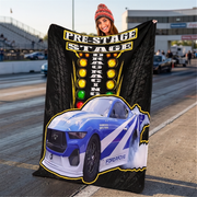 A person stands on a racetrack holding a large printed blanket or poster featuring a blue and white race car, tire graphics, and the words “PRE-STAGE DRAG RACING.” The racetrack and other people are blurred in the background.