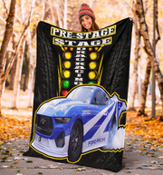 A person outdoors in a forest with fallen leaves holds up a large printed blanket or tapestry featuring a blue and white racing car, tire graphics, and the words 'PRE-STAGE DRAG RACING'.