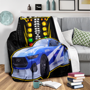 A printed throw blanket draped over a gray sofa in a living room setting. The blanket features a blue and white drag racing Ford Mustang with a large vertical racing light panel graphic on a black background.