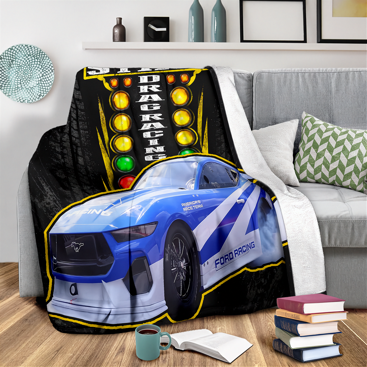 A printed throw blanket draped over a gray sofa in a living room setting. The blanket features a blue and white drag racing Ford Mustang with a large vertical racing light panel graphic on a black background.