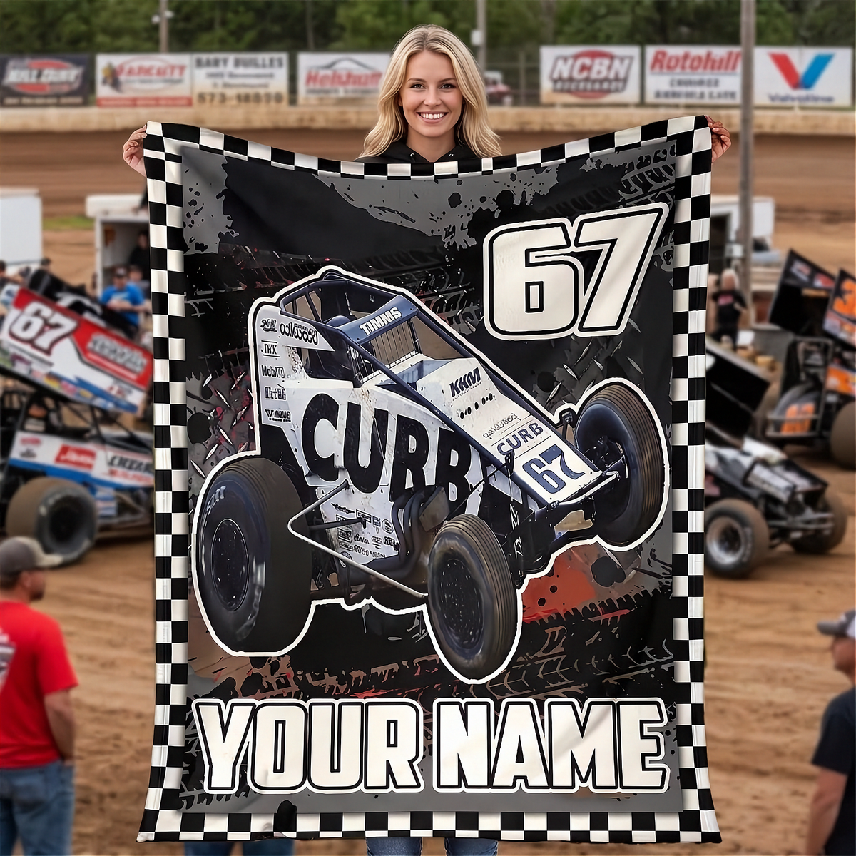 A woman stands on a dirt racetrack holding up a large custom-printed blanket or poster featuring a white and black NON WING race car with the number 67, a black-and-white checkered border, and the text 'YOUR NAME' at the bottom. Race cars and track signage are visible in the background.
