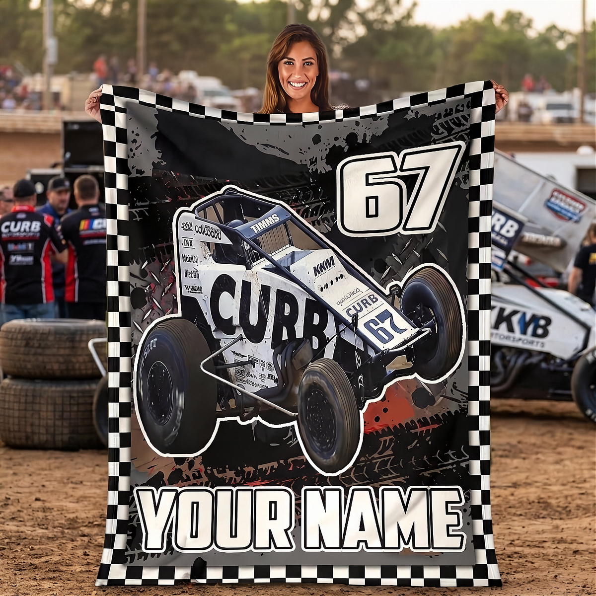 A woman stands in a dirt racing arena holding up a large custom-printed blanket or towel featuring a white and black NON-Wing race car with the number 67, a checkered border, and the text 'YOUR NAME'.