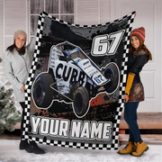 Two women standing in front of a white garage door holding up a custom fleece blanket featuring a white and black Non wing sprint race car with the number 67, a checkered border, and the text 'YOUR NAME'.