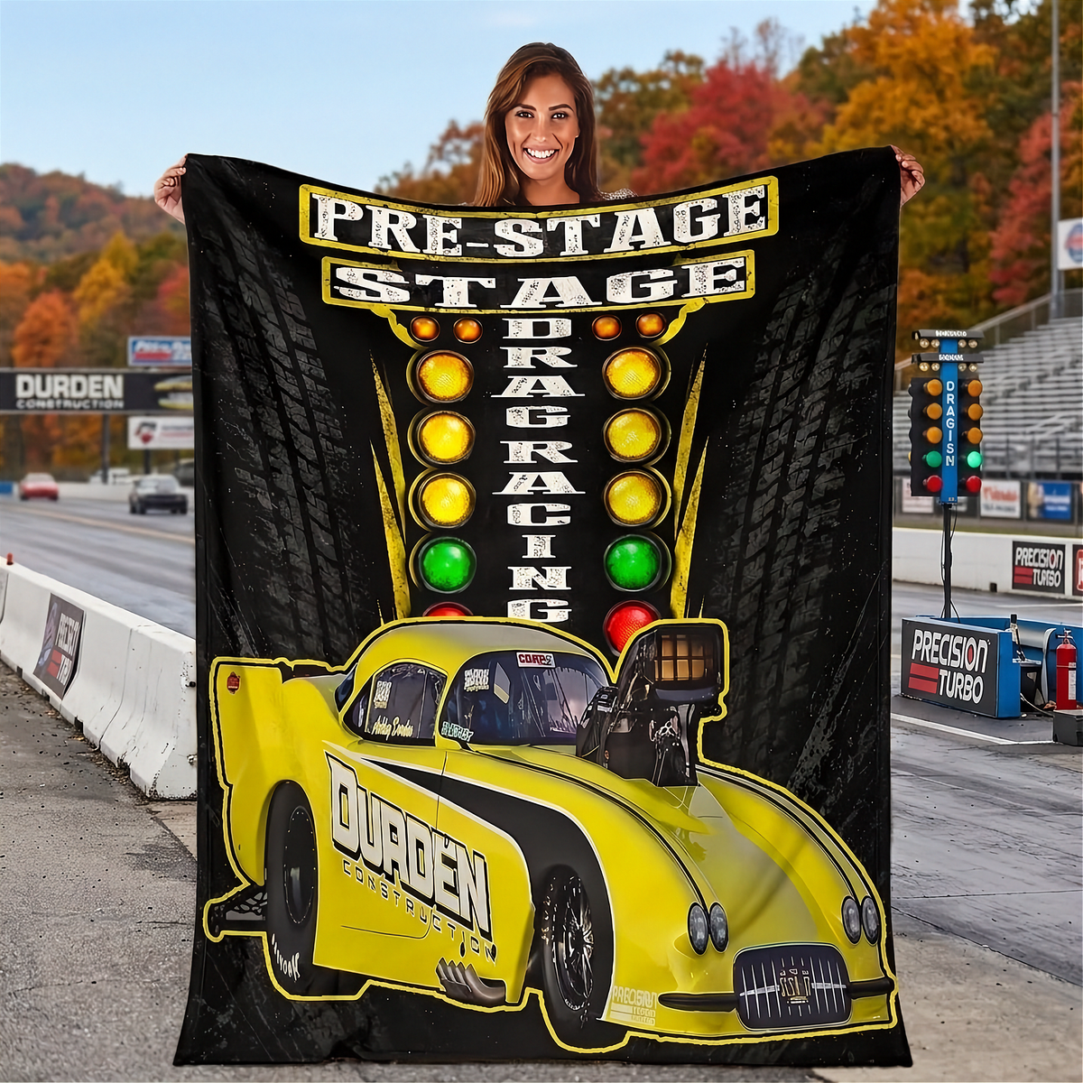 A woman stands on a racetrack holding up a large black fleece blanket or throw featuring a bright yellow drag racing car and the words 'PRE-STAGE DRAG RACING.' The background shows the racetrack, trees with autumn foliage, and race signage.