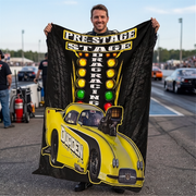 A man stands on a racetrack holding up a large black and yellow drag racing themed blanket or poster. The design features the words 'PRE-STAGE STAGE DRAG RACING' above a bright yellow drag car with 'DODGEN' on the side, and a vertical column of illuminated race lights.