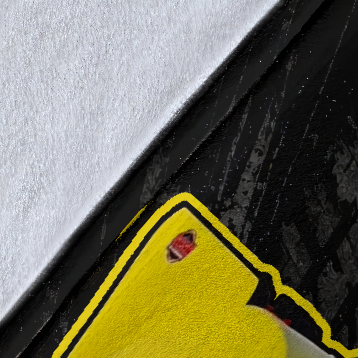 Close-up detail of a bright yellow sticker or decal with a black outline, placed over a black textured surface with a white textured area at the top.